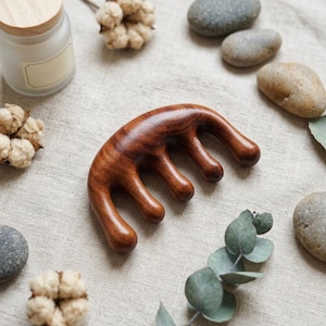 Könnte beinhalten: Ein hölzerner Kopfhautmassager mit gebogenem Griff und abgerundeten Zinken. Das Massagegerät besteht aus dunkelbraunem Holz und ist von dekorativen Elementen wie Steinen, Watteknospen und einer Kerze umgeben.