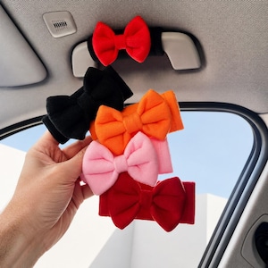 May include: A collection of five fabric bow accessories in red, black, orange, pink, and dark red. Each bow is attached to a matching fabric band. One red bow is mounted on a car grab handle, while the others are held in a stack for display.