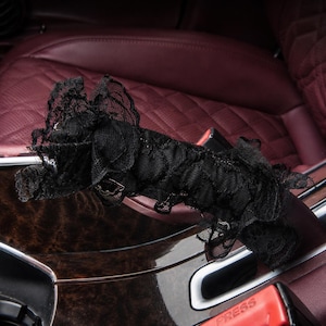 May include: A black lace cover featuring a ruffled, layered texture designed to wrap around a car seat belt buckle. The fabric is sheer with a delicate floral pattern, and the word PRESS is visible on the red plastic buckle underneath.