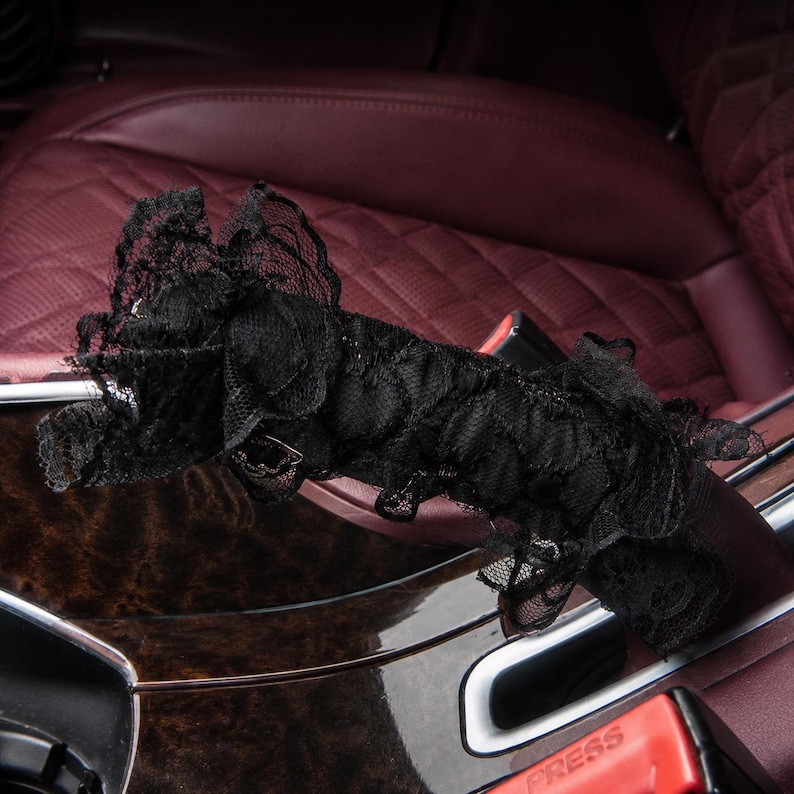 May include: A black lace cover featuring a ruffled, layered texture designed to wrap around a car seat belt buckle. The fabric is sheer with a delicate floral pattern, and the word PRESS is visible on the red plastic buckle underneath.