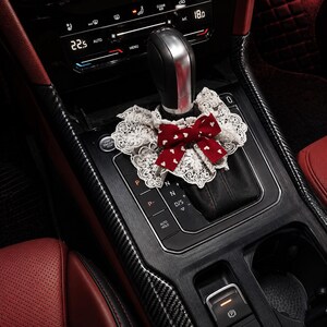 May include: A decorative accessory for a car gear shifter featuring a white lace ruffled collar and a dark red fabric bow with a small white heart pattern. The accessory is positioned around the base of a black and silver gear shift lever.
