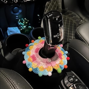 May include: A circular, multi-colored crochet cover featuring a scalloped edge with small, attached pom-poms in various colors. The piece is secured around the base of a black car gear shifter with a single white button closure at the front.