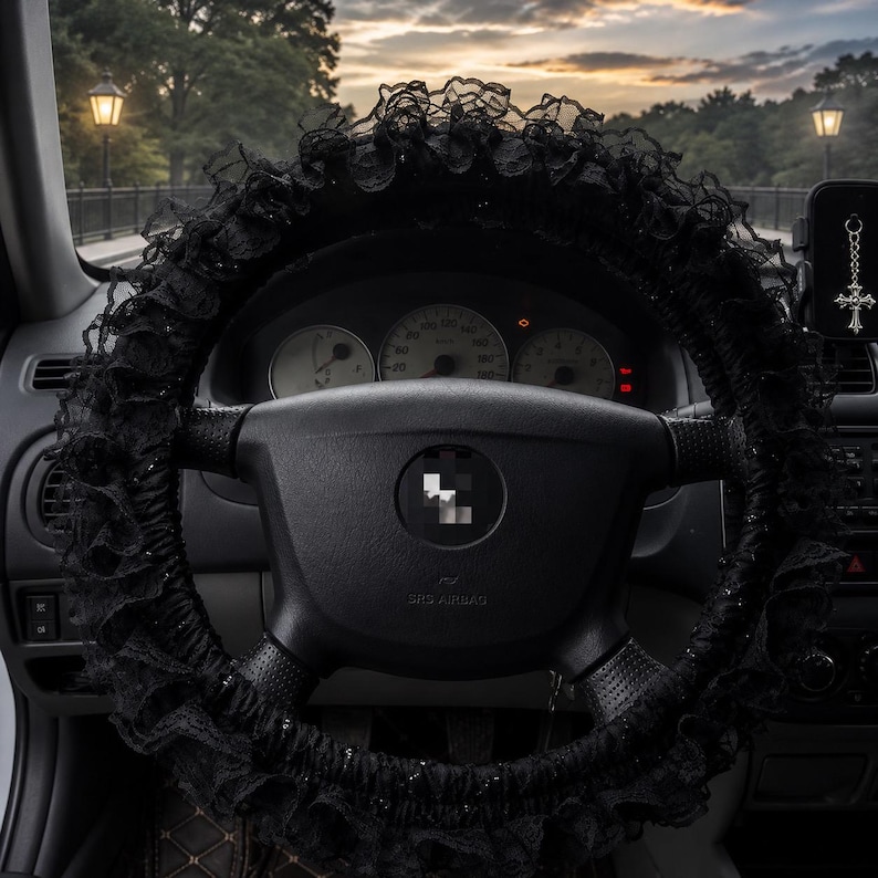 May include: A black steering wheel cover featuring ruffled lace trim and subtle glitter accents, installed on a car steering wheel. The cover surrounds the central hub and spokes, providing a decorative texture to the interior of the vehicle.