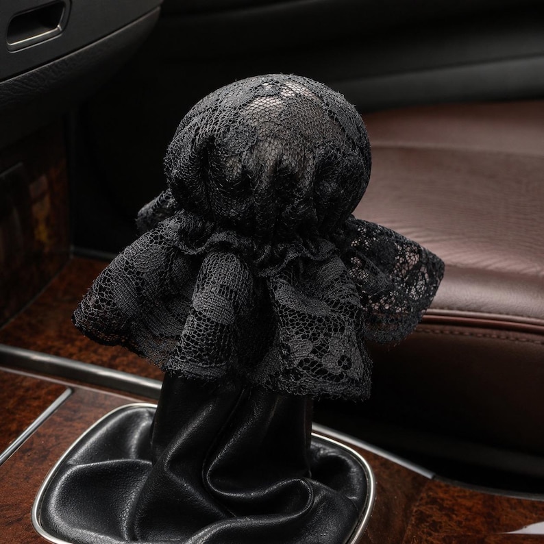 May include: A black lace gear shift knob cover featuring a rounded top and a ruffled skirt design. The cover is fitted over a black leather gear shift boot, creating a layered appearance with the intricate floral patterns of the lace fabric.