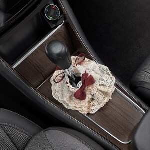 May include: A black car gear shift lever adorned with a decorative beige lace cover featuring a dark red ribbon bow. The lace fabric has a floral pattern and is gathered around the base of the gear stick, which is set in a dark wood grain console.