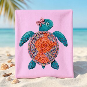 May include: A pink beach towel featuring a teal turtle illustration with an orange and yellow shell. The turtle has a pink bow and is set against a beach scene with sand and seashells.