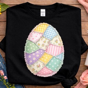 May include: Black t-shirt featuring a patchwork Easter egg design. The egg is composed of various fabric patterns, including polka dots, stripes, and floral prints, in pastel colors. The shirt is displayed on a wooden surface with floral and denim accents.