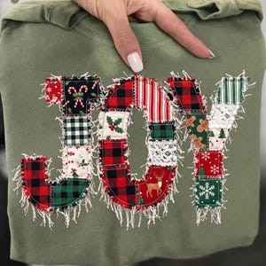 Può includere: Una felpa verde oliva con la parola JOY applicata in stile patchwork. Le lettere sono realizzate con vari tessuti a tema natalizio, tra cui motivi a quadri rossi e neri, bastoncini di zucchero, agrifoglio, renne e fiocchi di neve.