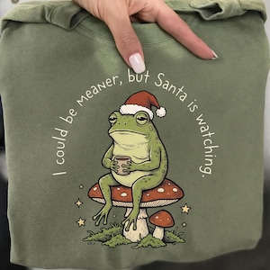 May include: An olive green sweatshirt featuring a graphic of a green frog wearing a red and white Santa hat, sitting on a mushroom while holding a small cup. The text "I could be meaner, but Santa is watching" is printed in an arc around the frog.