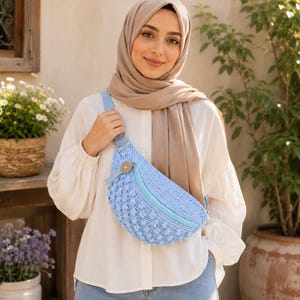 May include: A light blue crochet crossbody bag with a textured bobble stitch pattern, a light blue zipper closure, and a single wooden button accent. The bag features a matching light blue strap and is worn across the chest over a white long-sleeve shirt.