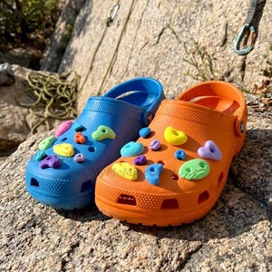 May include: A pair of blue and orange clogs, each adorned with colorful climbing-hold charms. The shoes are placed on a rock, with climbing gear visible in the background. The clogs have the word "MeMi" on the side.