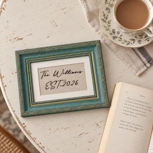 May include: A rectangular picture frame with a distressed teal finish and gold inner trim. The frame contains a white mat and a beige insert with the black text "The Williams EST. 2026" written in a cursive script font centered within the frame.