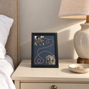 May include: A black rectangular picture frame with a dark blue mount containing two square black and white photographs. A red and white twisted cord connects the two photos, forming a continuous looping shape across the blue background.