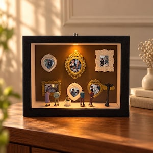 May include: A rectangular black shadow box display case featuring an internal light, a brown back panel, and five ornate miniature frames containing photographs. Small figurines of people stand on the base alongside a miniature street lamp.