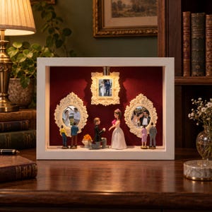 May include: A white rectangular shadow box frame with a deep red velvet backing, featuring three ornate cream-colored picture frames containing wedding photos, illuminated by a small top light, with miniature figurines of couples displayed below.