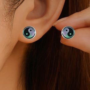 May include: A pair of round stud earrings featuring a yin and yang symbol design. The circular earrings have a metallic silver border surrounding a smooth, glossy surface with a gradient of light purple and teal colors forming the yin and yang pattern.