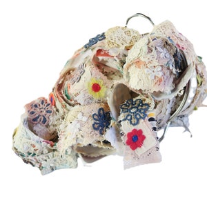 Puede incluir: Un arreglo decorativo de retazos de tela y encaje en un anillo de metal. Los textiles presentan una mezcla de colores, incluyendo crema, rosa y azul, con bordados y apliques florales. El efecto general es texturizado y en capas.