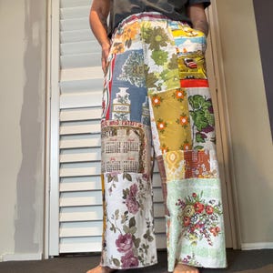 May include: Wide-leg trousers featuring a patchwork design made from various vintage-style fabrics. The patterns include floral prints, a 1979 calendar, a savoury spice label, and a landscape scene. The trousers have an elastic waistband and side pockets.