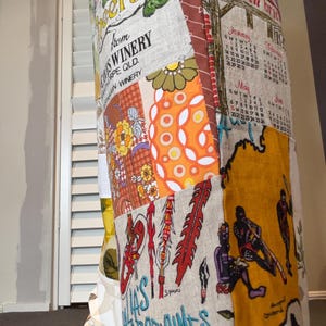 May include: A patchwork fabric item featuring various printed panels, including a winery label, a calendar, and illustrations of figures with spears. The textile displays a mix of orange, yellow, green, and brown colours with floral and graphic patterns.