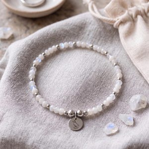 May include: A beaded bracelet featuring small, translucent white moonstone beads interspersed with tiny silver-toned metal spheres. A circular silver-toned metal charm engraved with a stylised fox silhouette hangs from the centre of the bracelet.