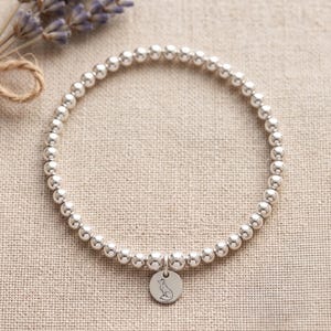 Può includere: Bracciale con perline d'argento con un piccolo ciondolo rotondo con l'illustrazione di una volpe. Il bracciale è composto da piccole perline rotonde d'argento. Lavanda e spago sullo sfondo.