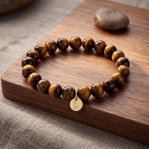 Op de afbeelding: Een kralenarmband gemaakt van gepolijste bruine en gouden tijgeroog edelstenen. De armband is voorzien van een ronde, goudkleurige metalen bedel die in het midden hangt, gegraveerd met een eenvoudige lijntekening van een zittende vos.
