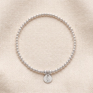 May include: A silver beaded stretch bracelet featuring a small, round, flat charm. The charm is engraved with a simple line drawing of a fox sitting in profile. The bracelet is composed of numerous small, polished, reflective silver-toned beads.