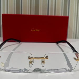 Puede incluir: Gafas sin montura con detalles dorados y patillas negras. Las gafas están colocadas frente a una caja roja con el logotipo de Cartier. Las lentes son claras y rectangulares.
