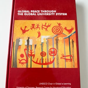 Peut inclure: Un livre à couverture rigide rouge intitulé "GLOBAL PEACE THROUGH THE GLOBAL UNIVERSITY SYSTEM". La couverture présente une illustration colorée avec des symboles rouges, jaunes et oranges. Le titre et les noms des auteurs sont imprimés en blanc. Publié par l'Université de Tampere.
