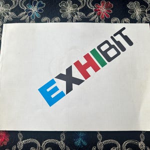Könnte beinhalten: Ein rechteckiges weißes Papierschild mit dem diagonal aufgedruckten Wort EXHIBIT. Die Buchstaben sind in Blau, Schwarz, Rot, Grün und Schwarz gehalten, wobei ein dezentes Gittermuster auf der gesamten weißen Papieroberfläche zu sehen ist.