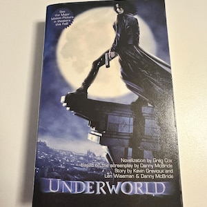 Può includere: Un libro tascabile intitolato "UNDERWORLD" con una copertina in stile manifesto cinematografico. La copertina presenta una donna con un lungo cappotto che tiene una pistola, sullo sfondo di una luna piena e di un paesaggio urbano. Il testo include "Vedi il film principale nei cinema questo autunno!"