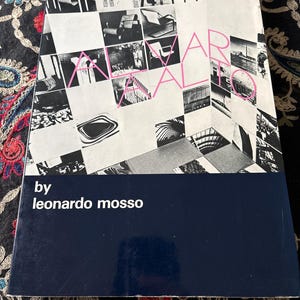 May include: A rectangular book with a white cover featuring a grid of black and white photographs of furniture and architecture. The text ALVAR AALTO is printed in pink, and the text by leonardo mosso is printed in white on a dark blue band.