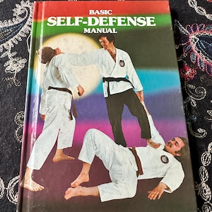 May include: A hardcover book titled Basic Self-Defense Manual. The cover features a graphic illustration of three individuals in white martial arts uniforms with black and brown belts, demonstrating self-defense techniques against a gradient background.