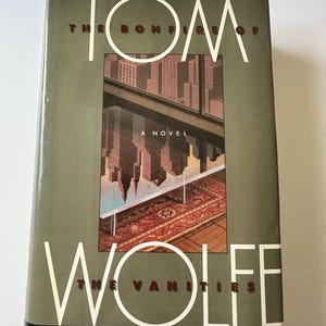 Puede incluir: Un libro de tapa dura titulado "The Bonfire of the Vanities" de Tom Wolfe. La portada presenta un paisaje urbano reflejado en una ventana, con el título y el nombre del autor en blanco y el título del libro en una fuente serif.