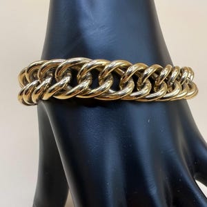 May include: A gold-toned chain link bracelet. The bracelet features a series of interlocking oval links, creating a bold and chunky design. The links are smooth and polished, reflecting light. The bracelet is displayed on a black surface.
