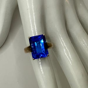 May include: A ring featuring a large, rectangular, faceted blue gemstone set in a metallic band. The stone is held in place by four small prongs at the corners, showcasing a vibrant, deep blue color with light reflections across the surface.