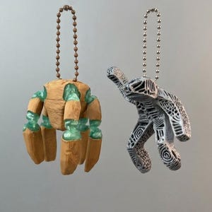 Pode incluir: Dois ornamentos em forma de mão. Um é castanho com detalhes verdes e uma corrente de bolas. O outro é cinzento com padrões intrincados e uma corrente de bolas prateada. Ambos estão pendurados num fundo neutro.