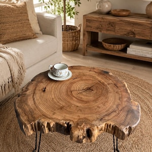 May include: A rustic, live-edge coffee table with a natural wood grain finish. The table is supported by black hairpin legs and sits on a woven jute rug. A teacup and saucer rest on the table's surface, suggesting a cozy living room setting.