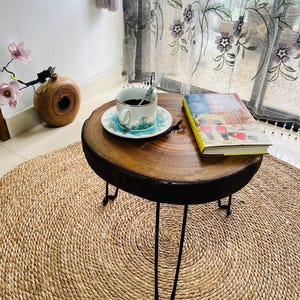 May include: A small, round wooden side table with black hairpin legs. A cup of coffee on a saucer sits on top, next to a book. The table is on a woven jute rug, creating a cozy, rustic aesthetic.