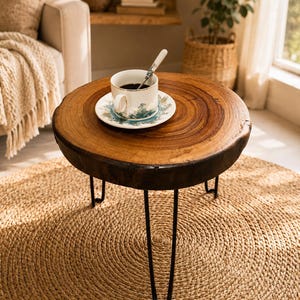 May include: A round side table featuring a natural wood slab top with visible grain and dark bark edges, supported by three slender black metal hairpin legs. A white ceramic cup and saucer with a blue and gold pattern sit on the table.
