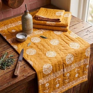 Könnte beinhalten: Senfgelber, gesteppter Tischläufer mit weißen Kreis- und Blumenmustern. Eine Keramikölflasche, eine kleine Schüssel und gefaltete Servietten liegen auf einer Holzoberfläche. Ein Kupfertopf hängt im Hintergrund.