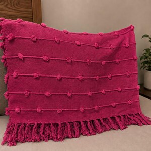 May include: A rectangular fuchsia throw pillow featuring horizontal rows of raised tufted pom-poms across the front surface. The bottom edge of the pillow is finished with a dense row of matching fuchsia fringe tassels hanging from the base.