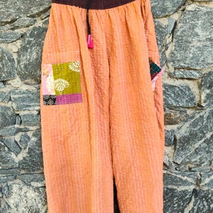 May include: Wide-leg pants in a light orange hue featuring vertical white stitching throughout. The garment has a dark brown elastic waistband with a black drawstring and a single patch pocket on the side with a floral pattern.