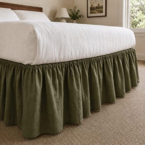 Può includere: Una balza per letto color verde oliva con un design arricciato e a volant che pende dalla base del letto. Il letto è coperto da un piumino bianco e cuscini bianchi, e la balza presenta una finitura in tessuto testurizzato fino al pavimento.