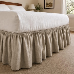 May include: A bed skirt made of light beige linen fabric with a gathered, ruffled design. The skirt is positioned around the base of a bed frame, featuring a textured weave and a consistent drop length that covers the lower portion of the bed.
