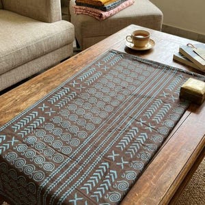 Pode incluir: Um caminho de mesa retangular castanho com um padrão repetitivo de motivos de espirais azul-claras e riscas geométricas em ziguezague. O tecido está estendido, exibindo uma impressão consistente em toda a superfície com linhas claras.