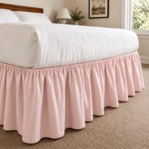 Puede incluir: Un faldón de cama de color rosa claro con un diseño fruncido y con volantes que se extiende desde la base del colchón hasta el suelo. La tela presenta una textura suave y mate y cubre la parte inferior del marco de la cama, creando capas.