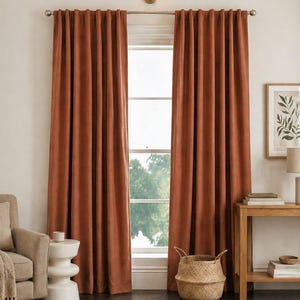 May include: A pair of floor-length, solid rust-coloured curtains hanging from a silver metal rod. The curtains feature a pleated header and a smooth, matte fabric texture, extending from the top of the window frame down to the floor.