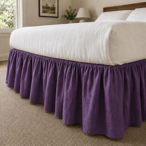 May include: A solid purple bed skirt with a gathered, ruffled design that hangs from the base of a bed. The bed is topped with a white mattress and white pillows, creating a contrast between the vibrant purple fabric and the neutral bedding.
