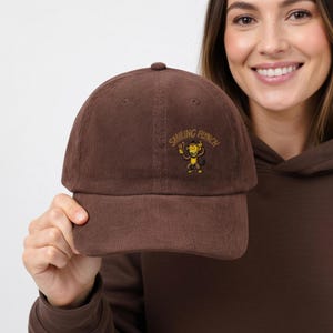 Puede incluir: Gorra de béisbol de pana marrón con el texto "Smiling Punch" y un gráfico de un mono de dibujos animados. La gorra tiene una visera curva y un botón cubierto de tela en la parte superior. El sombrero es de color marrón liso.
